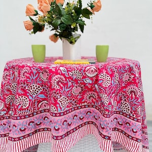 Puede incluir: Un mantel redondo fucsia con un estampado floral en blanco, azul y rosa. El mantel está sobre una mesa de mimbre blanca, con un jarrón de flores color melocotón y dos tazas verdes. El mantel tiene un borde a rayas.