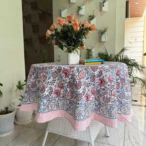 May include: A round tablecloth with a floral pattern in pink, blue, and white, on a white wicker table. The tablecloth has a pink and white striped border. A vase of peach roses sits on the table.