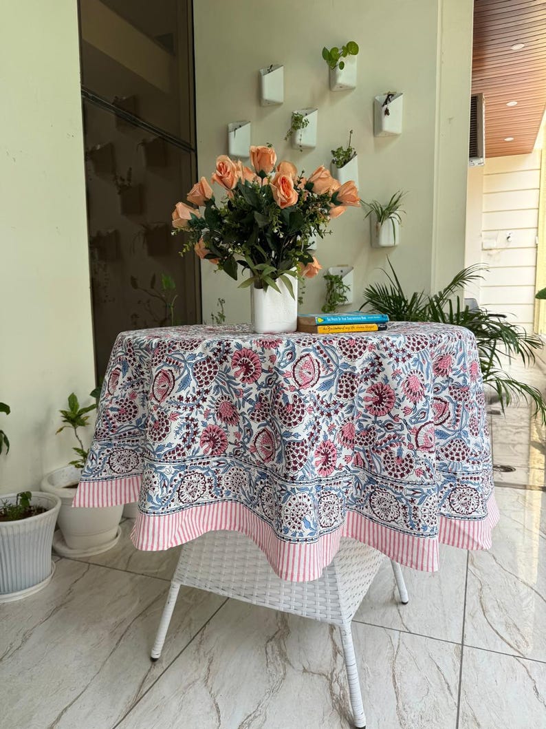 May include: A round tablecloth with a floral pattern in pink, blue, and white, on a white wicker table. The tablecloth has a pink and white striped border. A vase of peach roses sits on the table.