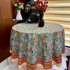 May include: A round tablecloth featuring a floral design in green, pink, yellow, and orange. The tablecloth is draped over a table, with a decorative border and striped design. A black guitar and vase with flowers are in the background.