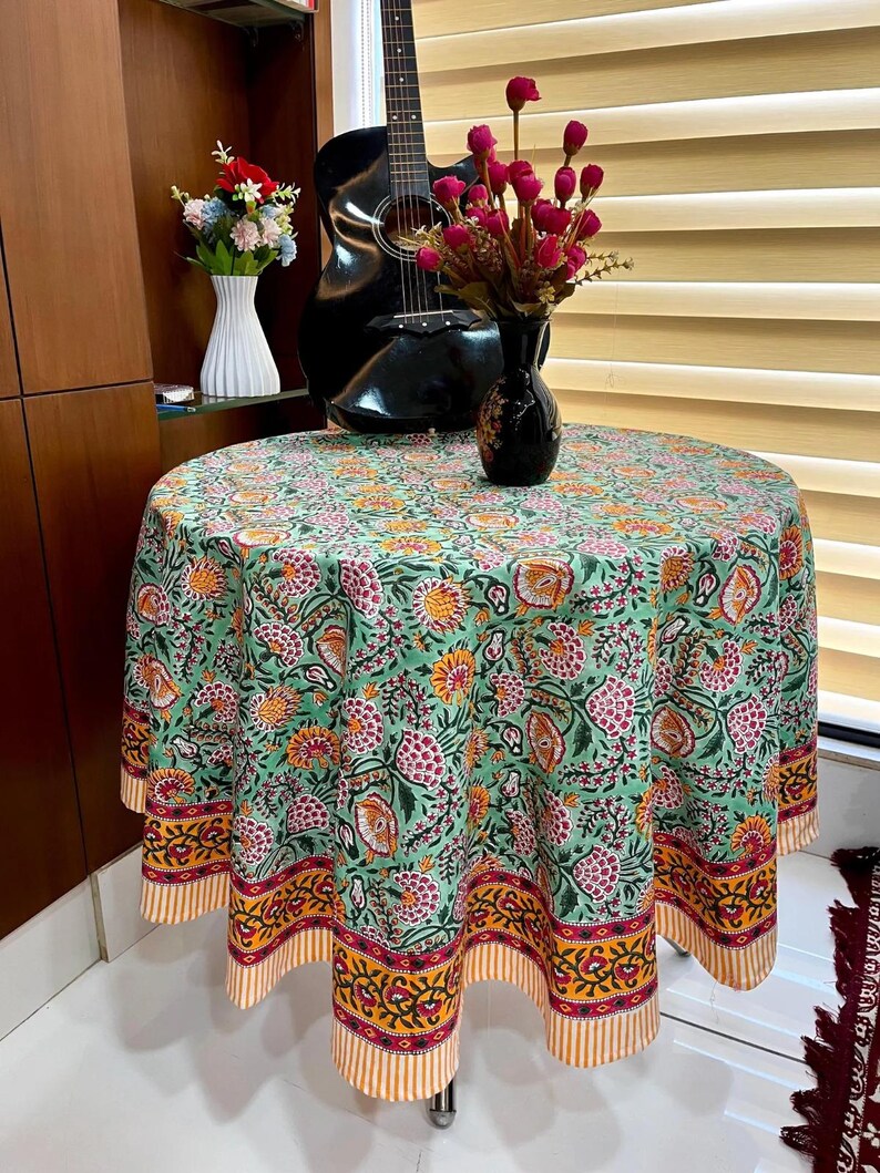 May include: A round tablecloth featuring a floral design in green, pink, yellow, and orange. The tablecloth is draped over a table, with a decorative border and striped design. A black guitar and vase with flowers are in the background.