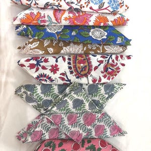 May include: A stack of eight folded fabric napkins featuring various block-printed floral and botanical patterns in colors including blue, orange, pink, brown, and grey on white backgrounds, arranged in a vertical column on a white surface.
