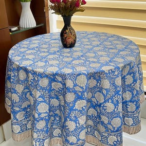 May include: A round blue tablecloth with a white floral design. The tablecloth has a brown and white striped border. A black vase with a floral design sits on the tablecloth. The tablecloth is on a round table.