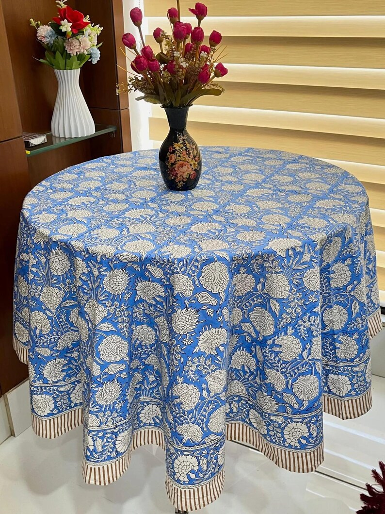 May include: A round blue tablecloth with a white floral design. The tablecloth has a brown and white striped border. A black vase with a floral design sits on the tablecloth. The tablecloth is on a round table.