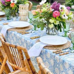 May include: A beautifully set table with a blue floral tablecloth, white plates, and woven placemats. Bamboo-style cutlery and green glassware complement the floral centerpieces. Wicker chairs add to the outdoor dining aesthetic.