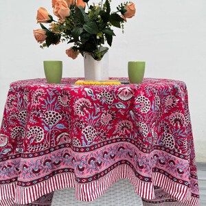 May include: A round tablecloth featuring a bold red floral design with pink and blue details. The table is decorated with a bouquet of peach roses in a white vase and two green cups. The tablecloth covers a white wicker table.