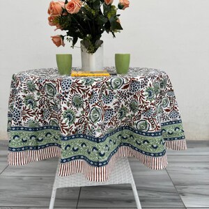 May include: A round tablecloth with a floral pattern in white, green, and brown covers a small white table. A bouquet of peach-coloured roses sits in a white vase. Two green cups and a book are on the table.