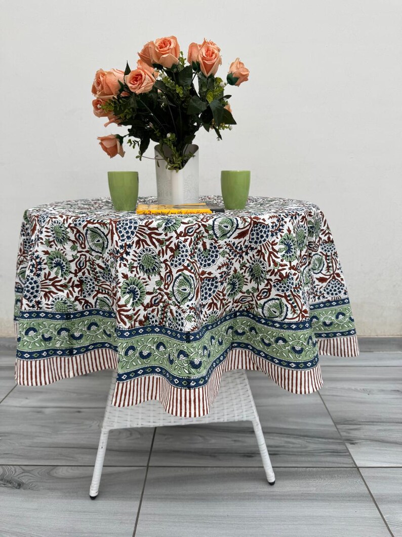 May include: A round tablecloth with a floral pattern in white, green, and brown covers a small white table. A bouquet of peach-coloured roses sits in a white vase. Two green cups and a book are on the table.