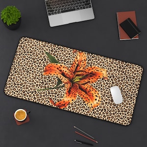 May include: Rectangular desk mat with a leopard print background and a large orange tiger lily. A white computer mouse sits to the right of the flower. The mat is surrounded by a black border.