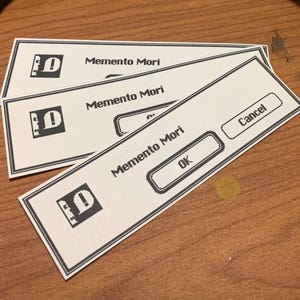 May include: A ripe banana with a yellow peel and green tips rests on a wooden surface. Beside it are three white rectangular cards with the text "Memento Mori" and a computer icon. One card has "OK" and "Cancel" buttons.