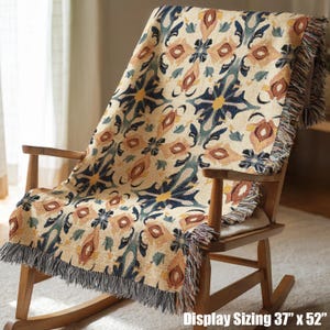 May include: A beige throw blanket with a repeating floral pattern in shades of blue, orange, and yellow is draped over a wooden rocking chair. The blanket has a fringed edge and the text "Display Sizing 37" x 52"" in the bottom right corner.