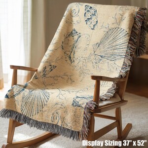 May include: A woven throw blanket featuring a beige background with a dark blue pattern of various seashells and coral branches. The blanket has fringed edges on the sides and bottom, and the text Display Sizing 37 inches by 52 inches.