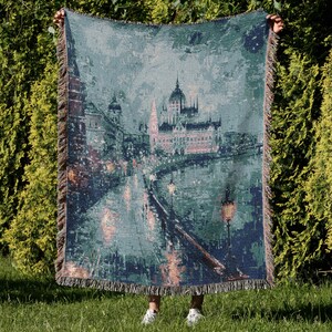 May include: A woven throw blanket featuring a cityscape scene with buildings, a river, and streetlights. The blanket has a teal, gray, and brown color palette with a fringed edge. The image depicts a European city at night.
