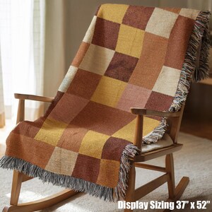May include: A woven throw blanket with a checkerboard pattern in shades of brown, tan, and yellow, draped over a wooden rocking chair. The blanket measures 37 x 52 inches and has fringed edges.