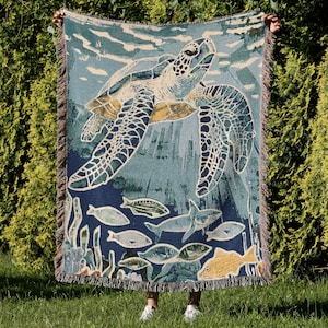 May include: Woven throw blanket with a sea turtle design, swimming amongst fish and coral. The blanket is in shades of blue, green, and beige, with a fringed edge. The design depicts an underwater scene.