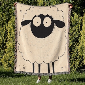 May include: A beige throw blanket featuring a large, whimsical sheep design. The sheep has a black face and legs, with a fluffy white body outlined in black. The blanket has a fringed edge and is held up against a green background.