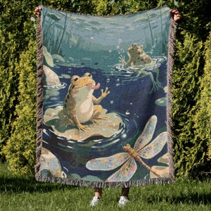 May include: A woven throw blanket featuring a pond scene with frogs, lily pads, and a dragonfly. The blanket has a dark blue background with green and beige accents. The edges are fringed.