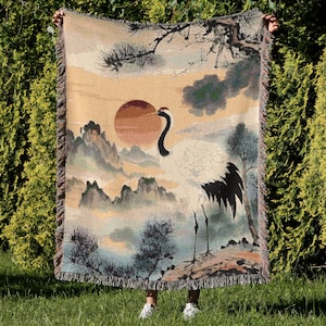 May include: A woven throw blanket featuring a crane standing in front of a sunset with mountains in the background. The blanket has a beige background with a detailed illustration in shades of orange, blue, and green. The edges are fringed.