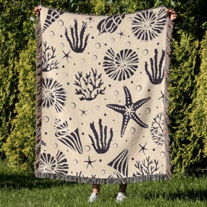 May include: A beige throw blanket with a repeating pattern of black seashells, starfish, and coral. The blanket has a fringed edge and is held up against a backdrop of green foliage. The design includes various marine life elements.