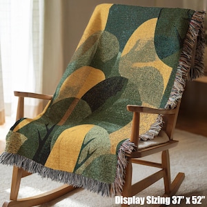 May include: A woven throw blanket featuring a repeating abstract pattern of dark green, olive, and mustard yellow shapes with black tree-like silhouettes. The blanket has fringed edges on the top and bottom and measures 37 inches by 52 inches.