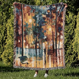 May include: A woven throw blanket featuring an impressionistic landscape scene with a park bench, streetlights, and a person walking. The color palette includes shades of brown, blue, and yellow. The blanket has a fringed edge.