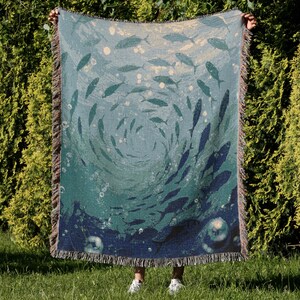 May include: A woven throw blanket featuring an underwater scene with a school of fish swimming in a circular pattern. The blanket has a teal and blue color scheme with a fringed edge. The design includes bubbles and a gradient effect.