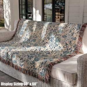 May include: A woven throw blanket with a cream background and a blue floral pattern is draped over a white wicker sofa. The blanket measures 50 x 60 inches and has fringed edges with red, brown, and cream colors. The text "Display Sizing 50" x 60"" is visible.