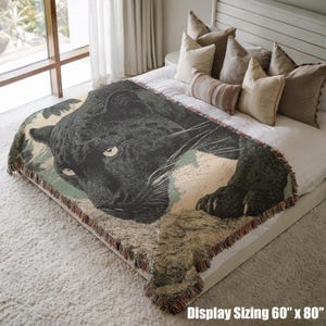 May include: A woven throw blanket featuring a black panther design. The blanket is rectangular, approximately 60 inches by 80 inches, with a fringed edge. The panther is depicted in a detailed, realistic style, with a neutral color palette.