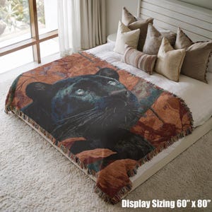 May include: A woven throw blanket featuring a black panther with blue eyes, set against a backdrop of autumn leaves. The blanket measures 60 inches by 80 inches and has a fringed edge. The design is displayed on a bed with pillows.
