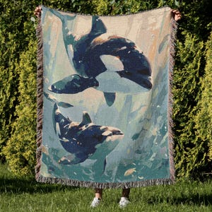 May include: Woven throw blanket featuring a marine scene with two orcas swimming in blue-green water. The blanket has a fringed edge and a beige background. The orcas are depicted in shades of blue and black.