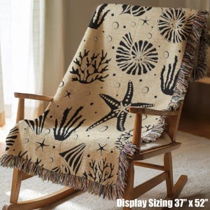 May include: A woven throw blanket featuring a tan background with a black pattern of seashells, starfish, and coral. The blanket has fringed edges and measures 37 inches by 52 inches. The design includes various marine life motifs scattered across.