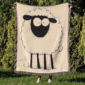 May include: Beige throw blanket featuring a whimsical black and white sheep illustration. The sheep has large eyes and a fluffy woolly body. The blanket has a fringed edge and is held up against a backdrop of green foliage.