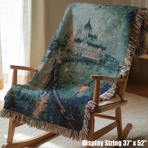 May include: A woven throw blanket draped over a wooden rocking chair. The blanket features a scenic design with a building and landscape in shades of green, blue, and beige. The blanket measures 37 x 52 inches.