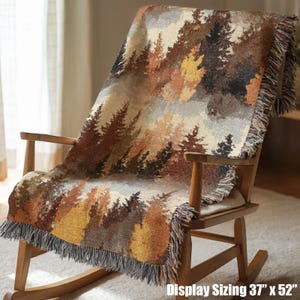 May include: A woven throw blanket featuring a forest landscape with pine trees in shades of brown, gold, and cream. The blanket has a fringed edge and measures 37 inches by 52 inches. It is draped over a wooden rocking chair with a smooth finish.