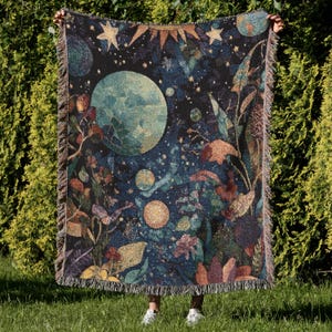 May include: A woven throw blanket featuring a celestial design with planets, stars, and botanical elements in shades of blue, green, and brown. The blanket has a fringed edge and is held up against a backdrop of green foliage.