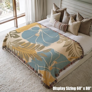 May include: A woven throw blanket with a floral design. The blanket features large blue, yellow, and green flowers and leaves on a cream background. The blanket measures 60 inches by 80 inches and has a fringed edge.