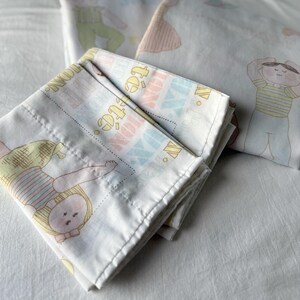 Op de afbeelding: Witte lakens met een herhaald patroon van poppen en het woord "TOYLAND" in pastelkleuren. De lakens zijn opgevouwen, met een deel van een tweede laken zichtbaar op de achtergrond, eveneens met poppenillustraties.