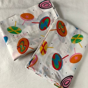 Puede incluir: Dos fundas de almohada blancas con un patrón de piruletas de colores. Las piruletas son rojas, amarillas, azules, verdes, naranjas y rosas. Las fundas de almohada tienen un fondo blanco con lunares negros.