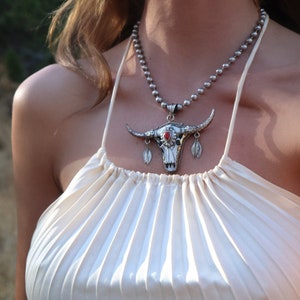 May include: A silver bull skull pendant necklace with a red stone in the eye. The necklace is on a woman wearing a white halter top.