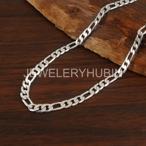 Può includere: Una collana a catena Figaro argentata su una superficie di legno. La catena presenta un motivo ripetuto di maglie allungate e circolari. Il gioiello è luminoso e riflettente, con il testo "JEWELERYHUBIN" visibile.