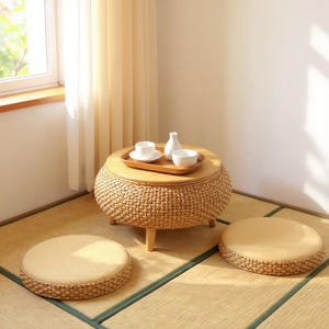 Op de afbeelding: Een ronde rotan salontafel met een houten blad, met een dienblad met witte kopjes en een vaas. Twee bijpassende ronde vloerkussens liggen op een tatami-mat. De kamer wordt verlicht door natuurlijk licht.