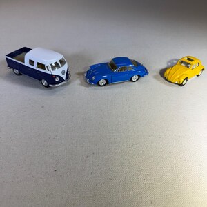 May include: Three miniature toy cars: a blue and white Volkswagen van, a blue sports car, and a yellow convertible. The vehicles are detailed with realistic features, including wheels and windows. The toys are likely made of die-cast metal or plastic.