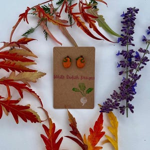 Puede incluir: Pendientes de botón con forma de calabaza naranja sobre una tarjeta marrón con el texto "White Radish Designs". Los pendientes están rodeados de colorido follaje otoñal y flores moradas, creando un tema de temporada.