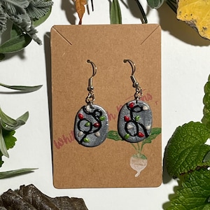 May include: A pair of silver-toned dangle earrings. Each earring features a gray oval with a black vine design and small red and white accents. The earrings are displayed on a brown card with the handwritten text "whimsical".
