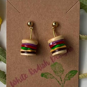 May include: A pair of novelty earrings shaped like miniature burgers. Each earring features a gold-colored post and a small, detailed burger with layers of bun, patty, lettuce, tomato, and cheese. The earrings are displayed on a brown card.
