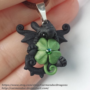 Op de afbeelding: Zwarte drakenhanger van polymer clay die een groen vierbladig klavertje vier met een groene strasssteen in het midden vasthoudt. De hanger is bevestigd aan een bruin koord.