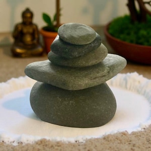 May include: A stack of smooth, gray river stones arranged in a balanced tower. The stones are on a white, sandy surface, with a blurred background featuring a small Buddha statue and a bonsai tree. The arrangement evokes a sense of peace and tranquility.