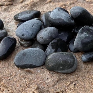 May include: A collection of smooth, rounded dark gray and black stones clustered together. The stones vary in size and shape, featuring polished, matte, and slightly damp textures that reflect light across their surfaces, creating a natural contrast.