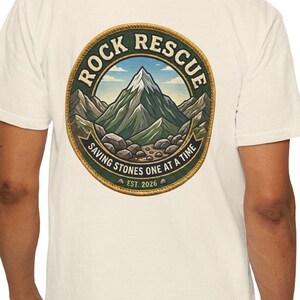以下が含まれることがあります： 背面に円形のグラフィックがプリントされたオフホワイトのTシャツ。デザインには山岳風景が描かれており、上部に「ROCK RESCUE」、中央に「SAVING STONES ONE AT A TIME」、下部に「EST. 2026」というテキストが縁取りの中に配置されています。