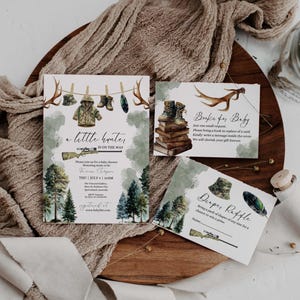 May include: Baby shower invitations and cards with a hunting theme. The main invitation features a watercolor illustration of a rifle, trees, and hunting gear. Additional cards include text "Books for Baby" and "Diaper Raffle".
